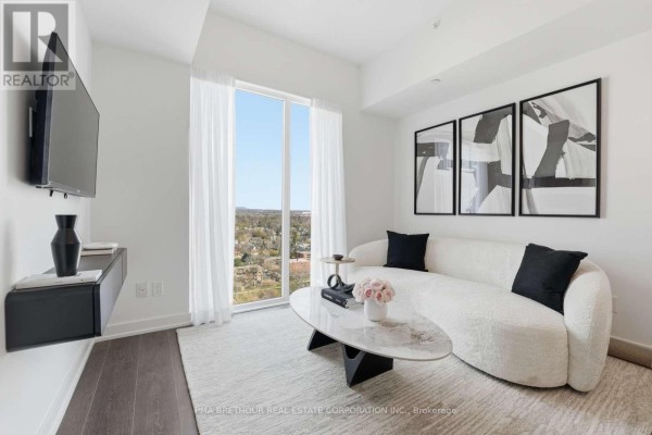 Living area featuring a white sofa, large window with curtains, and a mounted television. - 1901 - 370 Martha Street, Burlington