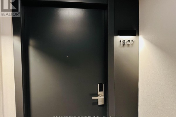 Close-up of a dark grey apartment door with a brushed nickel handle and the unit number 1907 illuminated above. - 1907 - 38 Gandhi Lane, Markham