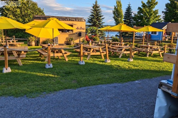 Wooden picnic tables and red chairs on a lawn with yellow umbrellas and service building. - 191 Water Street E, Prescott