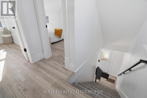 Hallway with wood-look floor, carpeted stairs, and black handrail leading to bedroom and exterior door. - 192 William Street W North Middlesex