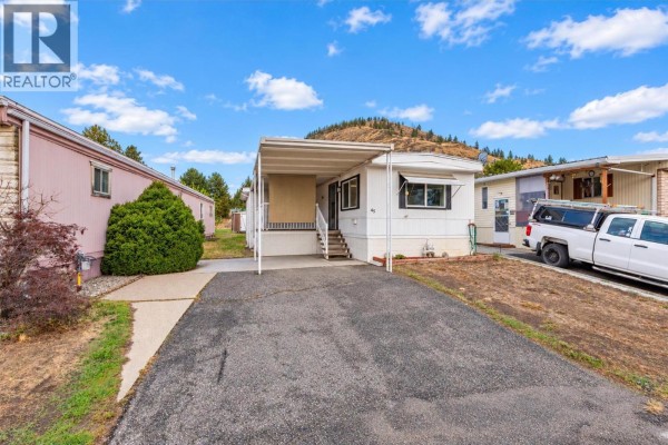Side exterior of a white mobile home with a covered carport, asphalt driveway, and adjacent pink mobile home. - 1929 97 Highway S Unit# 45, West Kelowna