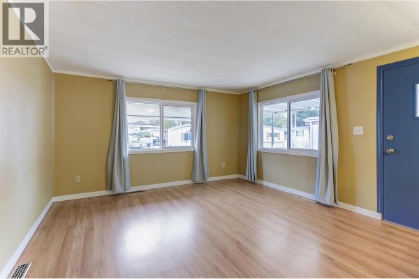 Living room with light wood laminate flooring, yellow walls, two windows with grey curtains, and a blue door. - 1929 97 Highway S Unit# 45, West Kelowna