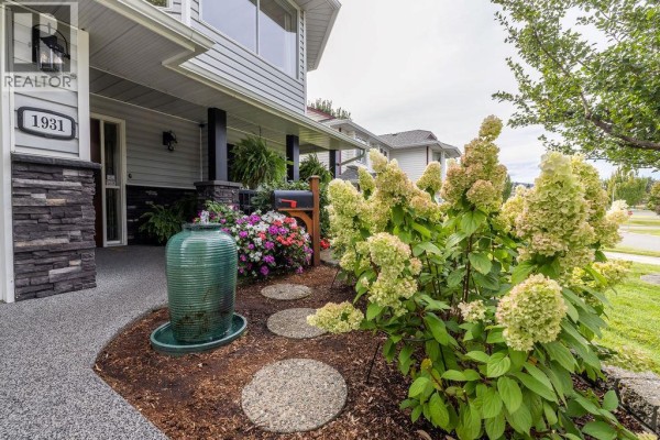 Close-up of the house entrance featuring stacked stone pillars, a green tiered water fountain, and a stepping stone path through flowering bushes. - 1931 Tinniswood Court, Kamloops