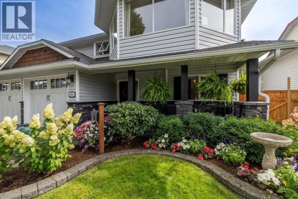 Exterior view of the house entrance showcasing a covered porch with hanging ferns, colorful flowerbeds, and a bird bath on the lawn. - 1931 Tinniswood Court, Kamloops
