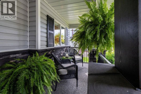 Covered porch with grey siding, black pillars, and wicker seating. Large hanging ferns create a lush, inviting atmosphere. - 1931 Tinniswood Court, Kamloops