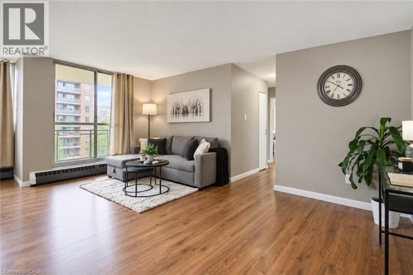 Living room with a grey sectional sofa, white rug, coffee table, and wood floors. - 1950 Main Street Unit# 705, Hamilton
