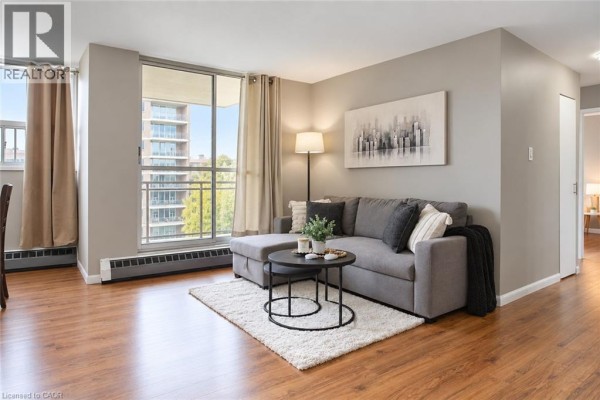 Living room featuring a sofa, rug, coffee table, and large window with balcony access. - 1950 Main Street Unit# 705, Hamilton