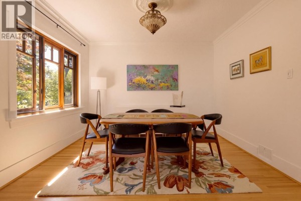 Dining room with a wooden table, chairs, floral rug, and a large window. - 1955 E 8th Avenue, Vancouver