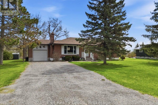Front view of brick bungalow with large pine trees and a gravel driveway. - 196 Elmwood Drive, Belleville