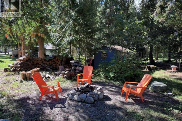 An outdoor fire pit with four orange Adirondack chairs, surrounded by stacked firewood and lush trees. - 1962 Tambellini Road, Christina Lake