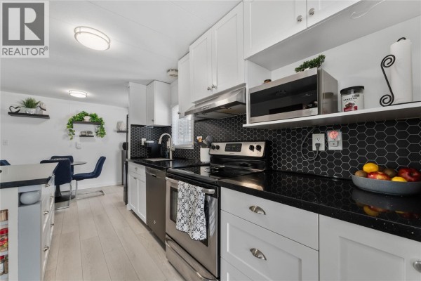 Modern kitchen with white cabinets, black quartz countertops, stainless steel appliances, and black hexagonal tile backsplash. - 1999 97 Highway S Unit# 85, West Kelowna