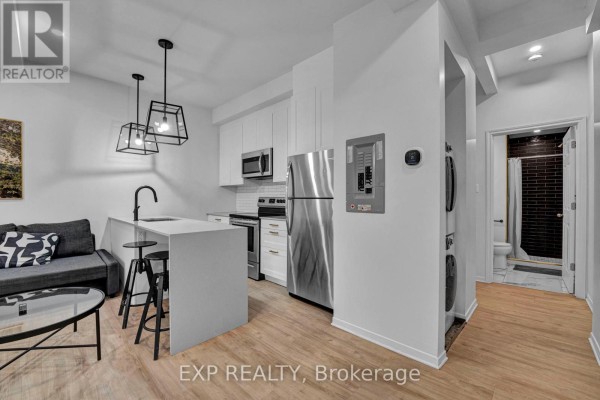 Combined living space with kitchen, grey sofa, stainless steel appliances, and view of laundry area. - 2 - 327 Catherine Street, Ottawa