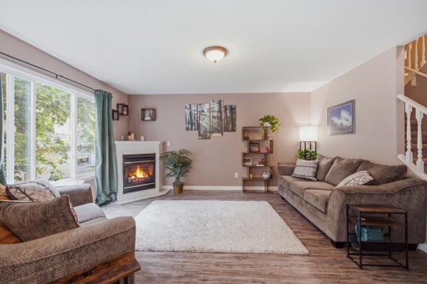 Living room with a fireplace, large window overlooking trees, sectional sofa, and a staircase with white railings. - 2 46360 Valleyview Road, Promontory, Chilliwack