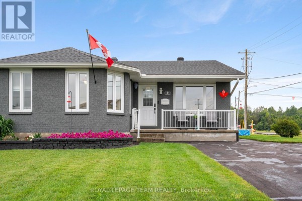 Front exterior of a grey brick bungalow with a white porch, railing, and Canadian flag. - 2 Billings Avenue W, South Dundas
