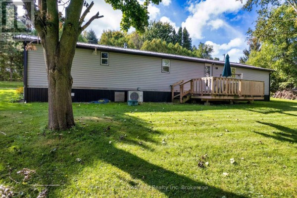 Backyard view of the grey mobile home with a wooden deck, green lawn, and mature trees. - 2 Snowbird Lane - 596101 59 Highway, East Zorra Tavistock
