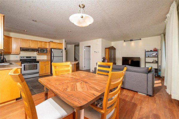 Dining area with wooden table and chairs, adjacent to kitchen and living room with television. - 20 Coulter Way, Brandon