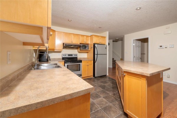 Kitchen with light wood cabinets, stainless steel appliances, island, and view of dining and living areas. - 20 Coulter Way, Brandon