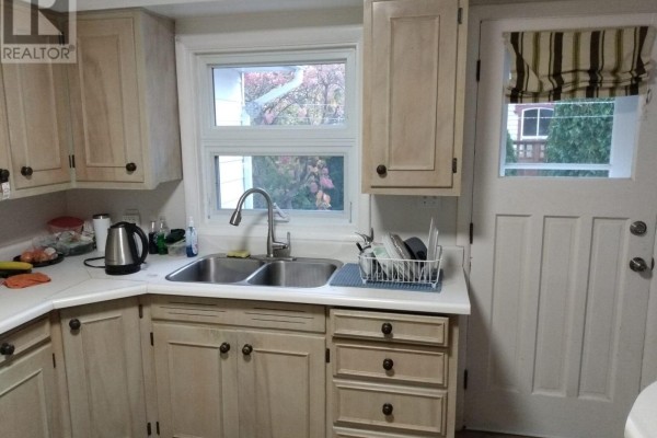 Compact kitchen with light wood cabinets, a double sink, faucet, kettle, and a door with striped curtains. - 20 Johnson Street, Prince Edward County
