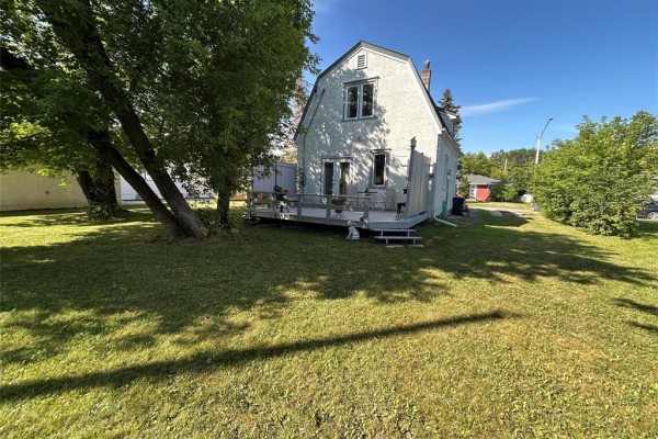 View of a white house with a large wooden deck in a grassy yard with trees. - 20 Poplar Street, Pine Falls