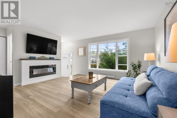 Living room with blue sofa, modern fireplace, large window with green trees view, and wood flooring. - 20 Stratford Drive, Cole Harbour