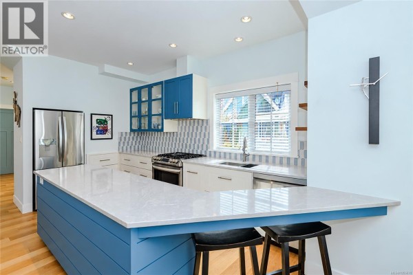 Kitchen with white cabinets, stainless steel appliances, a blue island with bar stools, and a window. - 200 Marina Way, Sooke