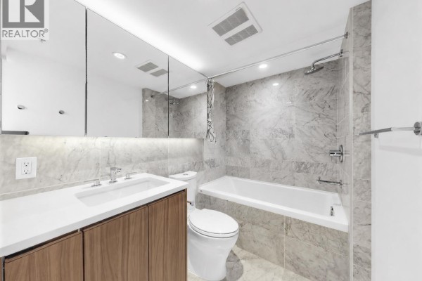 Bathroom with a white tub, toilet, vanity with wood cabinets, and marble tile walls. - 2007 433 Sw Marine Drive, Vancouver