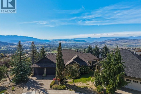 Aerial view of a large house with a three-car garage and surrounding trees against a mountainous backdrop. - 2009 Apex Court, Vernon