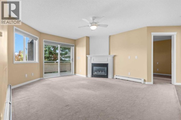Living room with beige carpet, white fireplace, and a ceiling fan. - 201, 20 Country Hills View Nw, Calgary