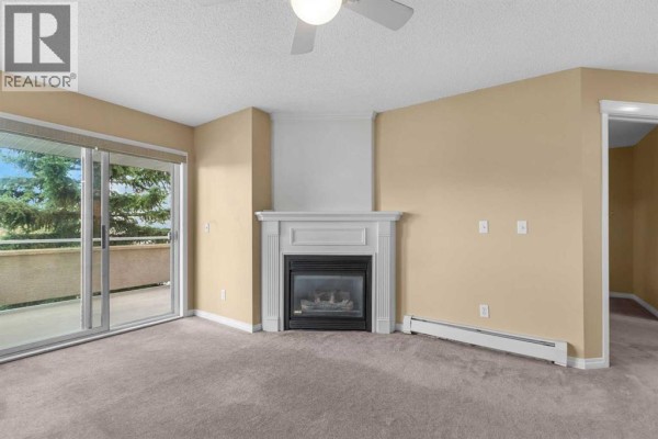 Living room with beige carpet, white fireplace, and glass doors opening to balcony. - 201, 20 Country Hills View Nw, Calgary