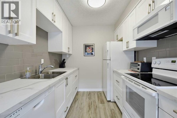 Galley-style kitchen with white cabinets, grey tile backsplash, and light wood flooring. - 201, 617 56 Avenue Sw, Calgary