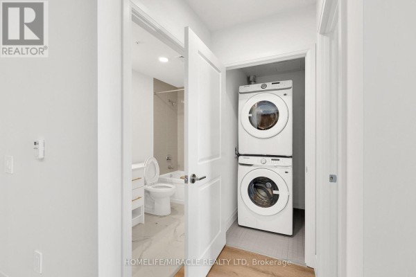 View of a stacked washer and dryer in a closet, with a bathroom containing a toilet and bathtub visible through an open door. - 201 - 1333 Weber Street E, Kitchener