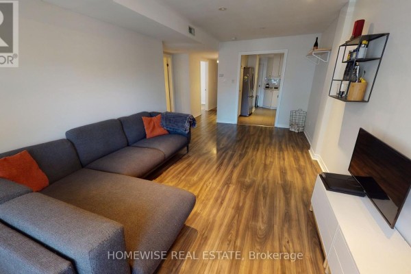 Living room facing a doorway to the kitchen, with a sectional sofa and wood-look flooring. - 201 - 145 Lansdowne Avenue, Toronto