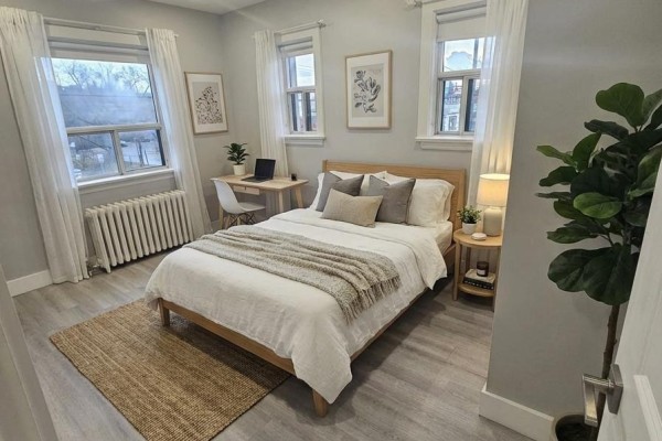 A bedroom with a queen bed, a small desk, white curtains, and a woven rug on the floor. - 201 - 1566 Avenue Road, Toronto