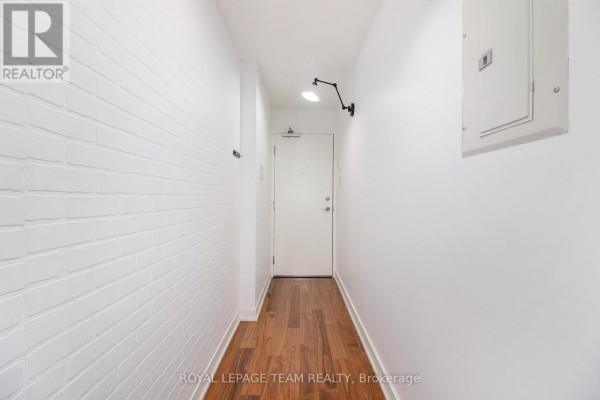 Apartment hallway with white brick accent wall, wood floor, and a single white door. - 201 - 354 Gladstone Avenue, Ottawa