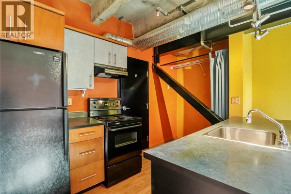 Interior view of an apartment with a metal door, stairs, kitchenette, and concrete pillar. - 201 1061 Fort St, Victoria
