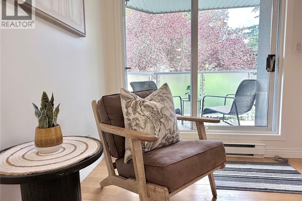 Armchair with decorative pillows next to a large sliding glass door with a view of trees. - 201 2520 Wark St, Victoria