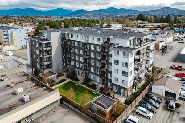 Aerial view of a modern apartment building surrounded by trees, parking, and distant mountains. - 201 32838 Ventura Avenue, Abbotsford