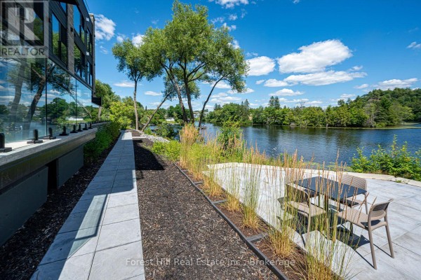 Exterior view of a modern building facade next to a patio and lake with lush greenery. - 202 - 32 Brunel Road S, Huntsville