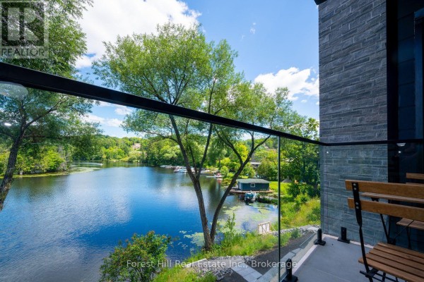 View of a calm lake and trees from a balcony with glass railings and outdoor seating. - 202 - 32 Brunel Road S, Huntsville