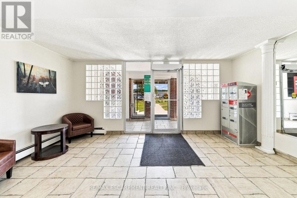 Apartment building lobby with tiled floor, glass block windows, mailboxes, and seating. - 202 - 851 Queenston Road, Hamilton