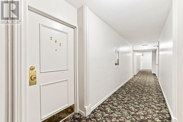 Hallway interior of apartment building with patterned carpet, white walls, and unit 202 door. - 202 - 851 Queenston Road, Hamilton