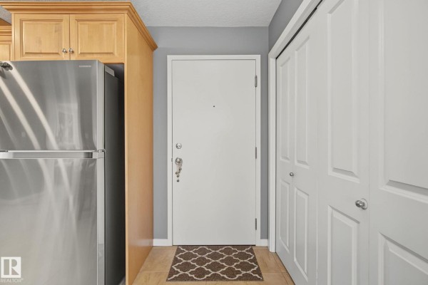 Apartment hallway with a white door, stainless steel refrigerator, and closet doors. - #202 11935 106st Nw, Edmonton