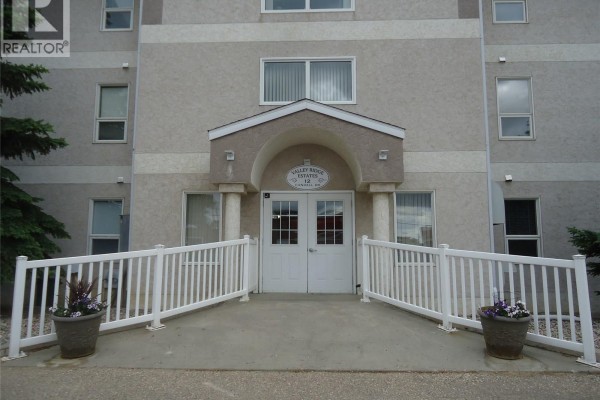 Front entrance of apartment building with double doors, covered archway, white railings, and potted flowers. - 202 12 Cundall Drive, Estevan