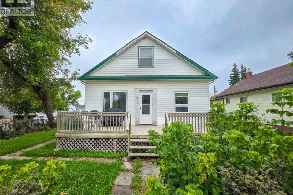 Front view of white siding house with green trim, wooden deck with furniture, and mature trees. - 202 6th Avenue E, Melville