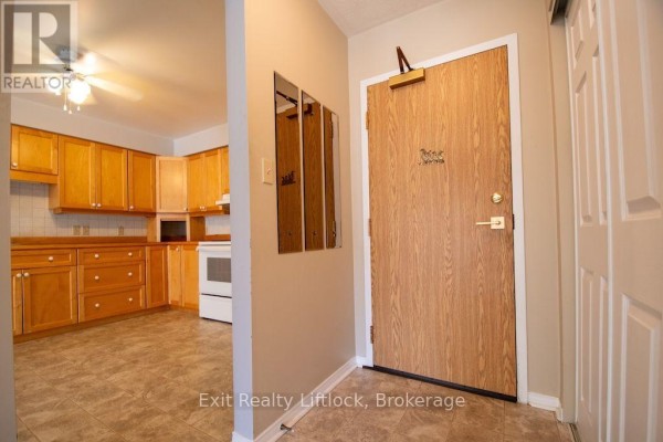 Apartment entrance door with wood grain finish and a small kitchen visible. - 202 C - 1099 Clonsilla Avenue, Peterborough