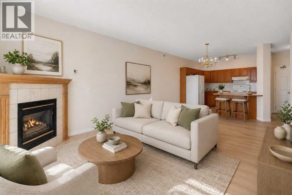 Living room with fireplace, sofa, coffee table, and kitchen in background. - 203, 4835 Dalhousie Drive Nw, Calgary