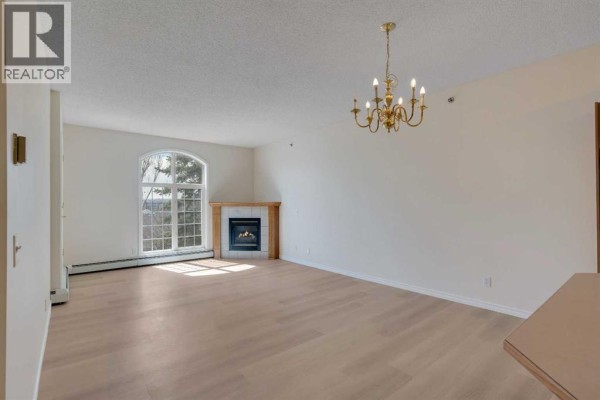 Living room with fireplace and arched window, dining area in background. - 203, 4835 Dalhousie Drive Nw, Calgary