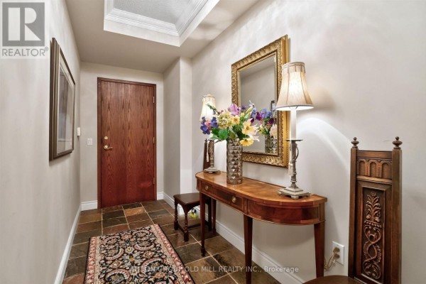 Foyer with wood door, console table, mirror, floral arrangement, and patterned rug. - 203 - 12 Old Mill Trail, Toronto