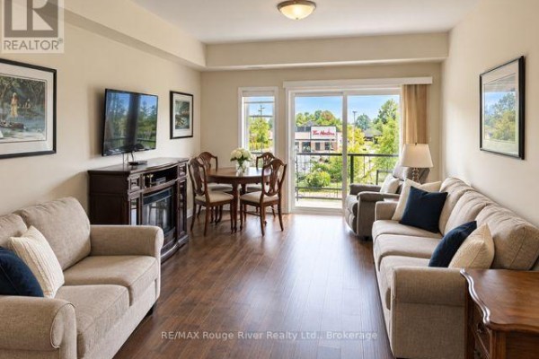 Living and dining area with beige sofas, dark wood furniture, and a sliding glass door to a balcony. - 203 - 340 Florence Drive, Peterborough