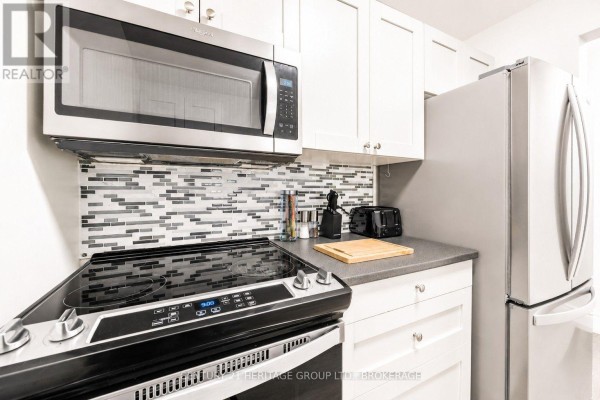 View of an apartment kitchen with light grey cabinets and a living room beyond. - 203 - 573 Armstrong Road, Kingston